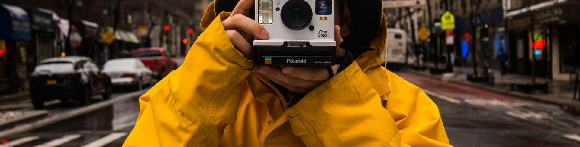 Mężczyzna w żółtej przeciwdeszczowej kurtce stoi na ulicy Nowego Jorku i trzyma aparat polaroid skierowany w stronę osoby robiącej zdjęcie.
