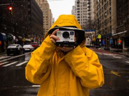 Mężczyzna w żółtej przeciwdeszczowej kurtce stoi na ulicy Nowego Jorku i trzyma aparat polaroid skierowany w stronę osoby robiącej zdjęcie.